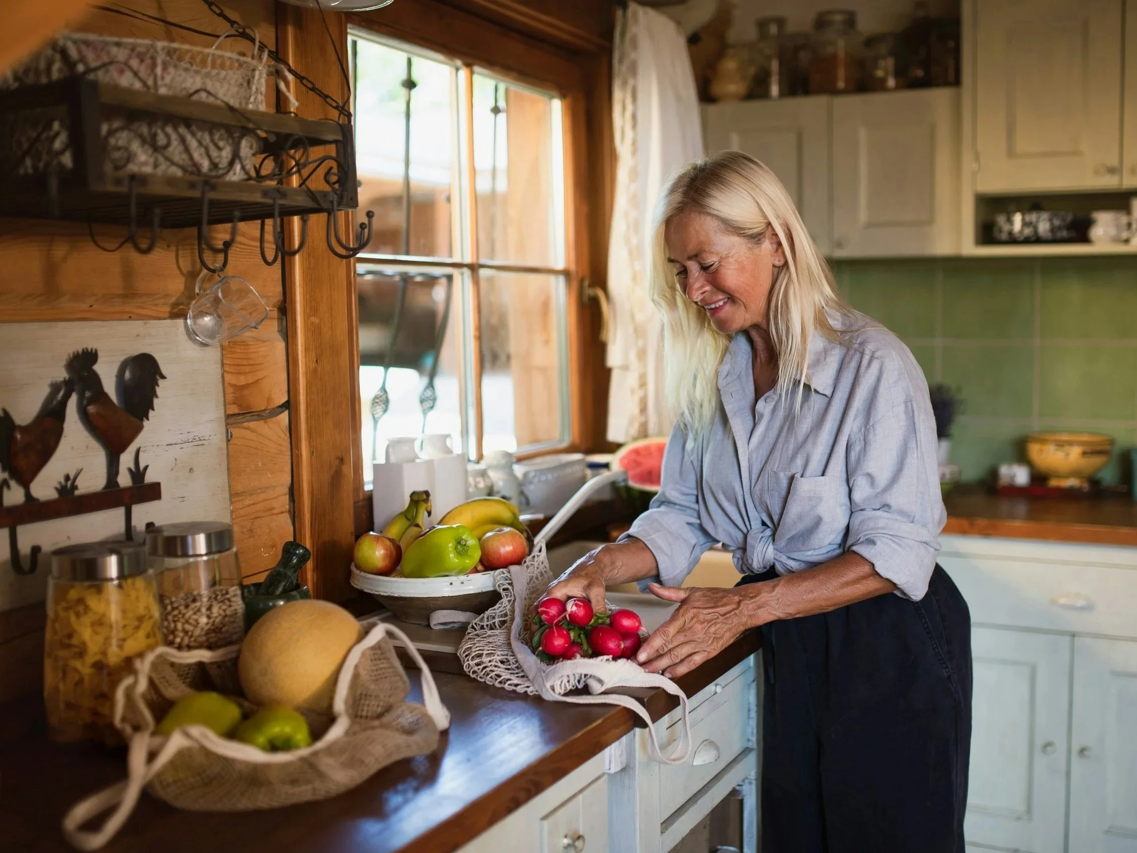 a woman smiles in her kitchen with her groceries, including a variety of fresh fruits and veggies.
