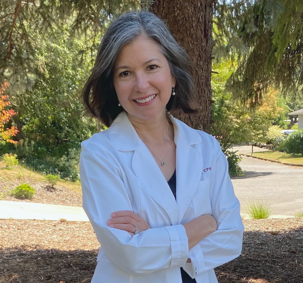 A female nurse practitioner stands in a white coat with her arms crossed, smiling, in front of trees.