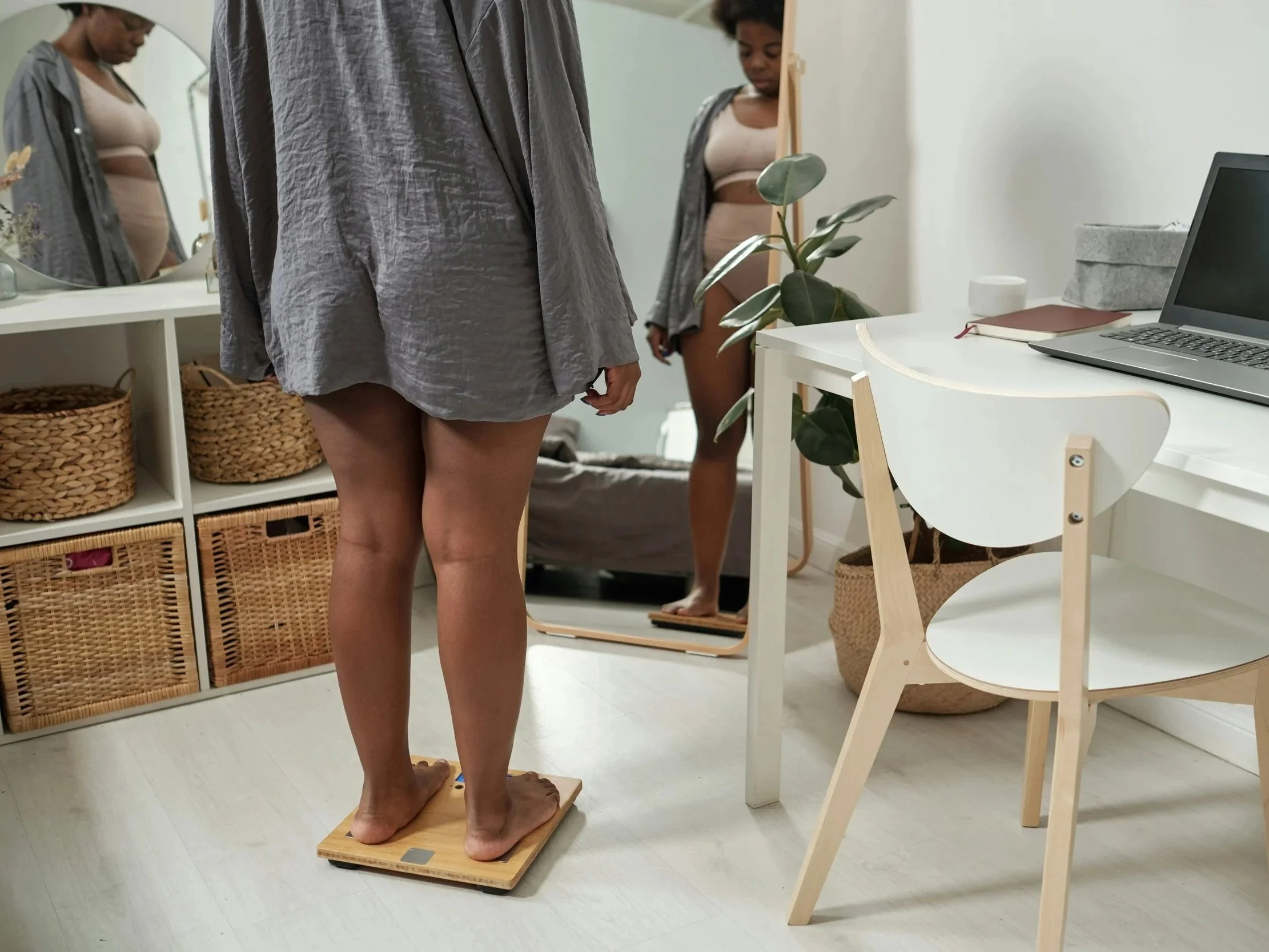 An overweight woman stands on a scale in front of a full length mirror. She looks down at the scale, not smiling, with her laptop open on the desk beside her.