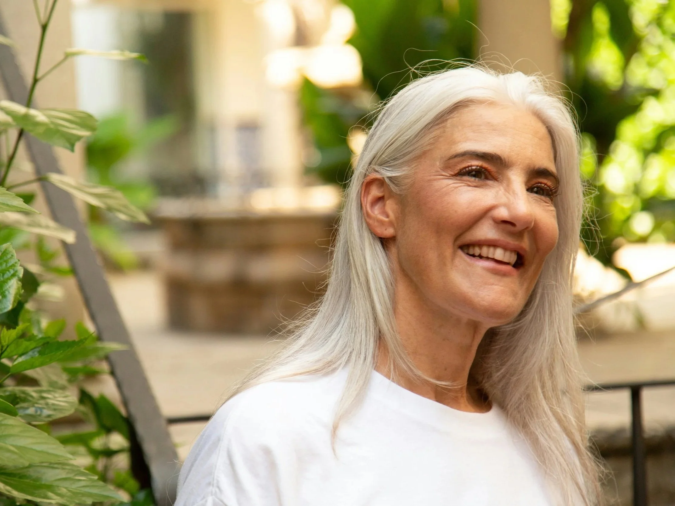 A healthy, mature, older woman with long, white hair smiles at something off camera. She is in a clean white shirt with greenery surrounding her.
