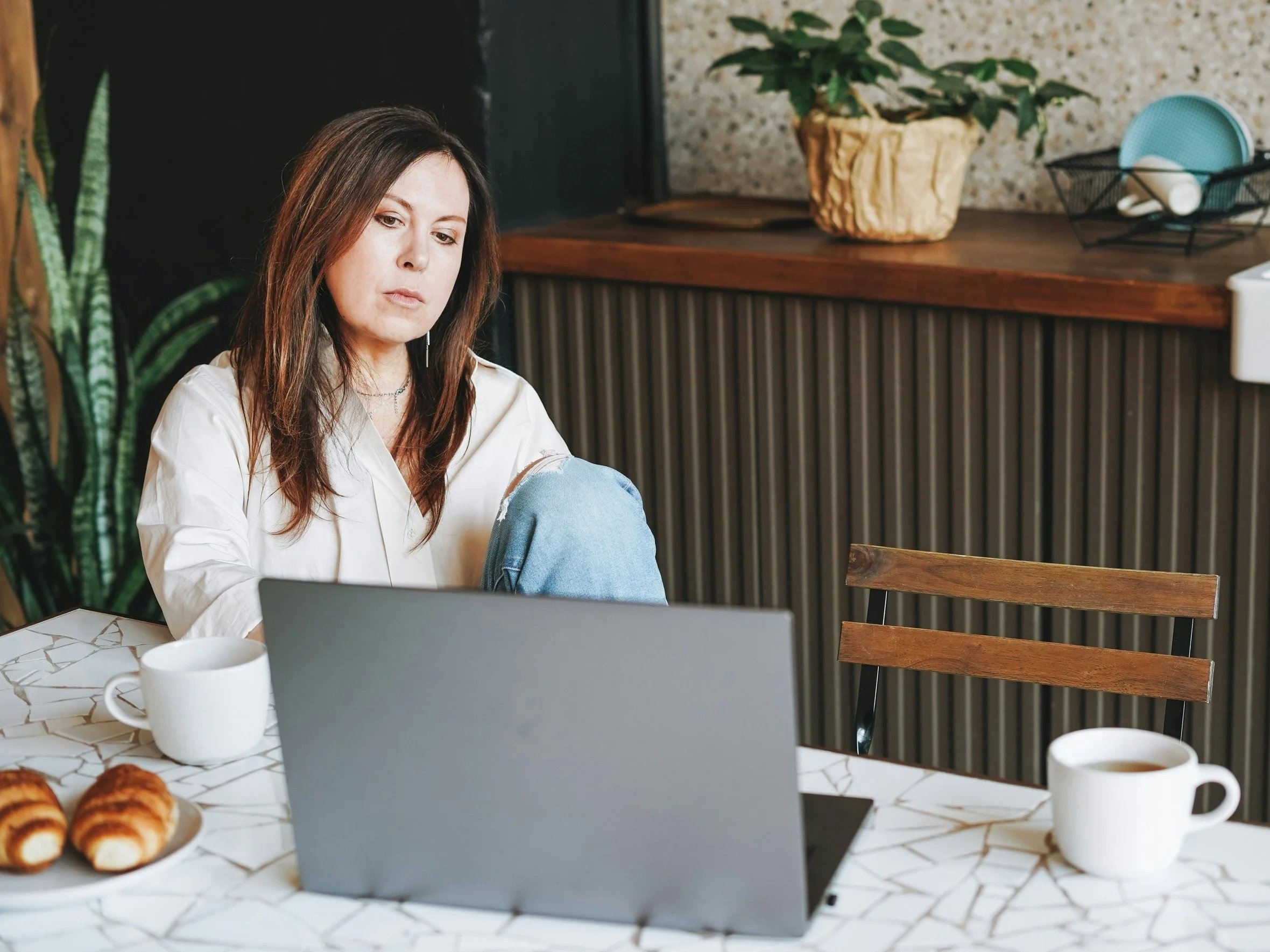A woman sits at her kitchen table in front of the computer, looking tired but not distressed, with two cups of coffee and a plate of croissants in front of her.