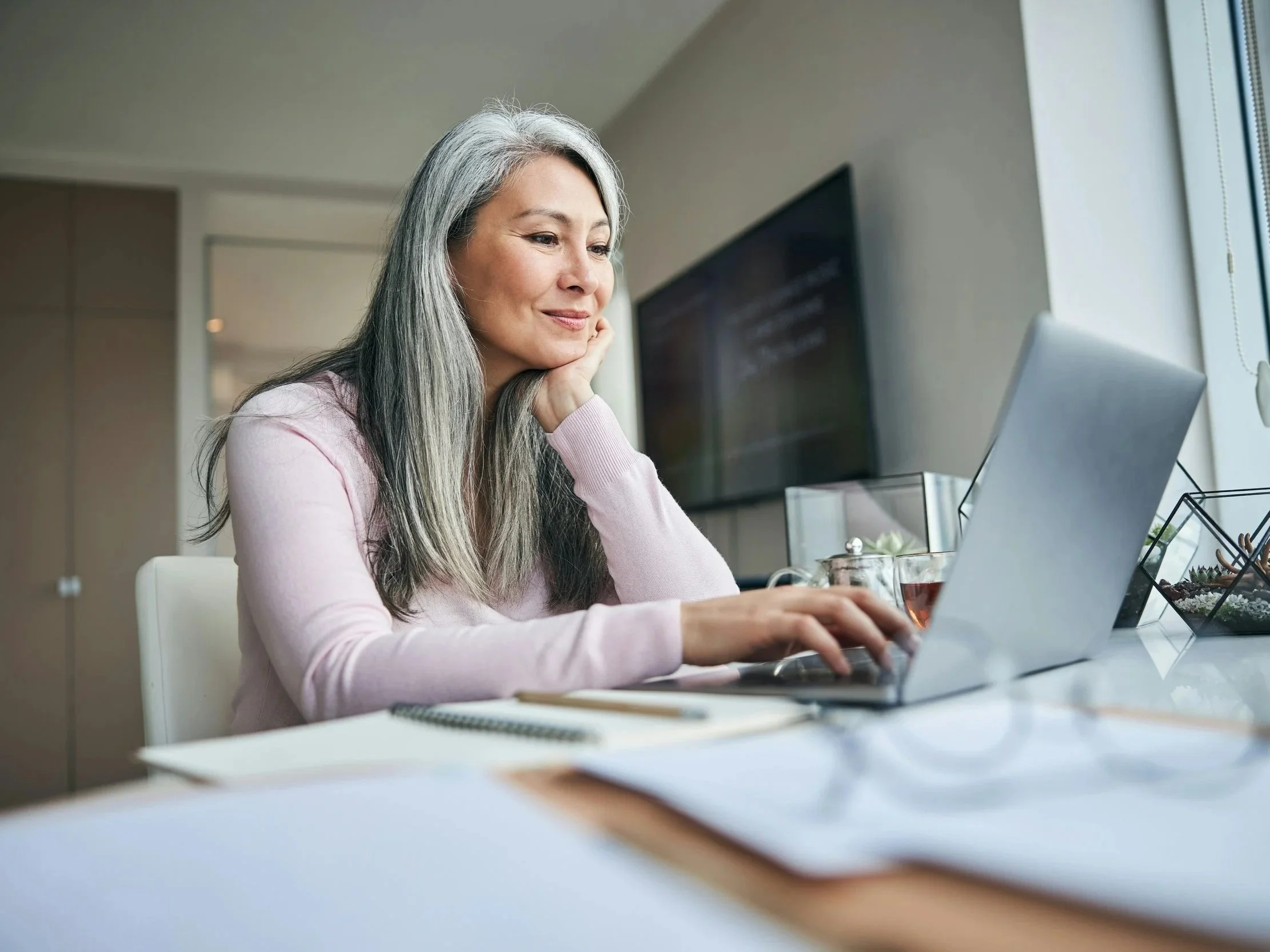 A woman smiles gently while working on her laptop with a cup of tea beside her.