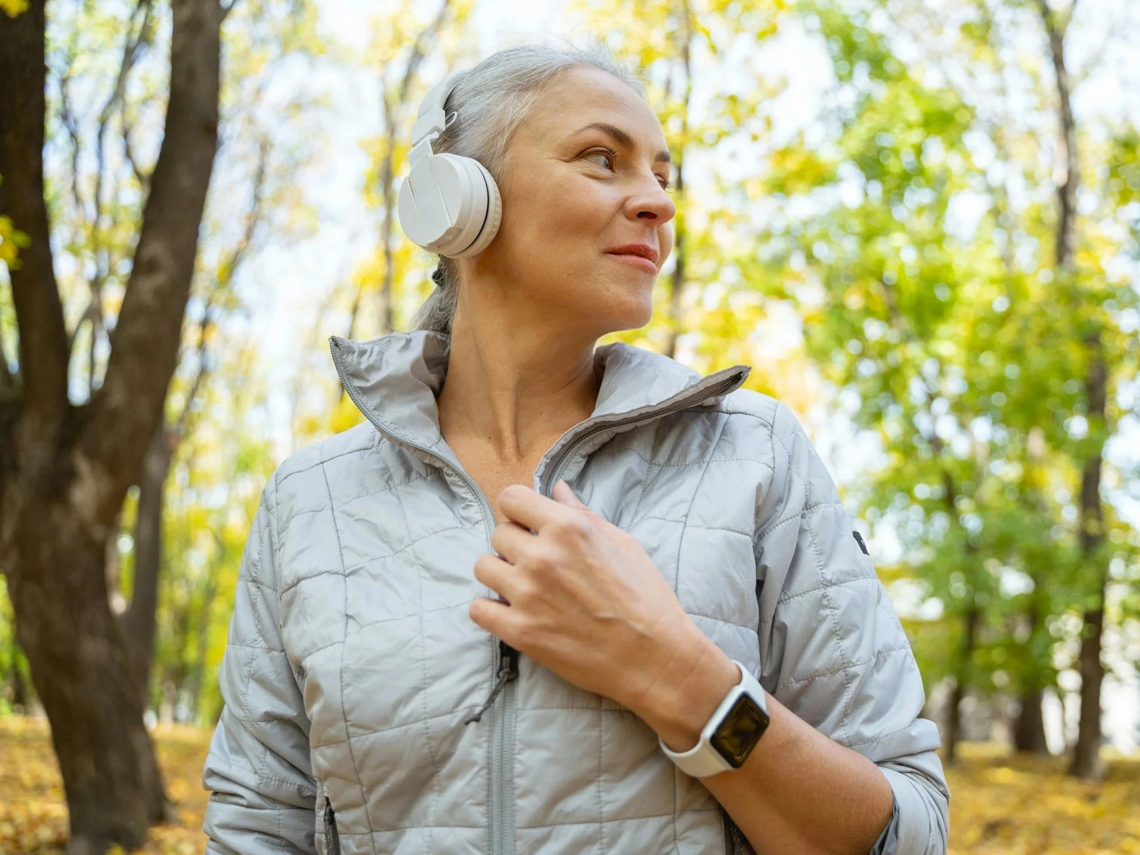 A woman stands in a wooded area, wearing a light jacket, headphones, and a smart watch. She gazes off to one side with a soft smile.