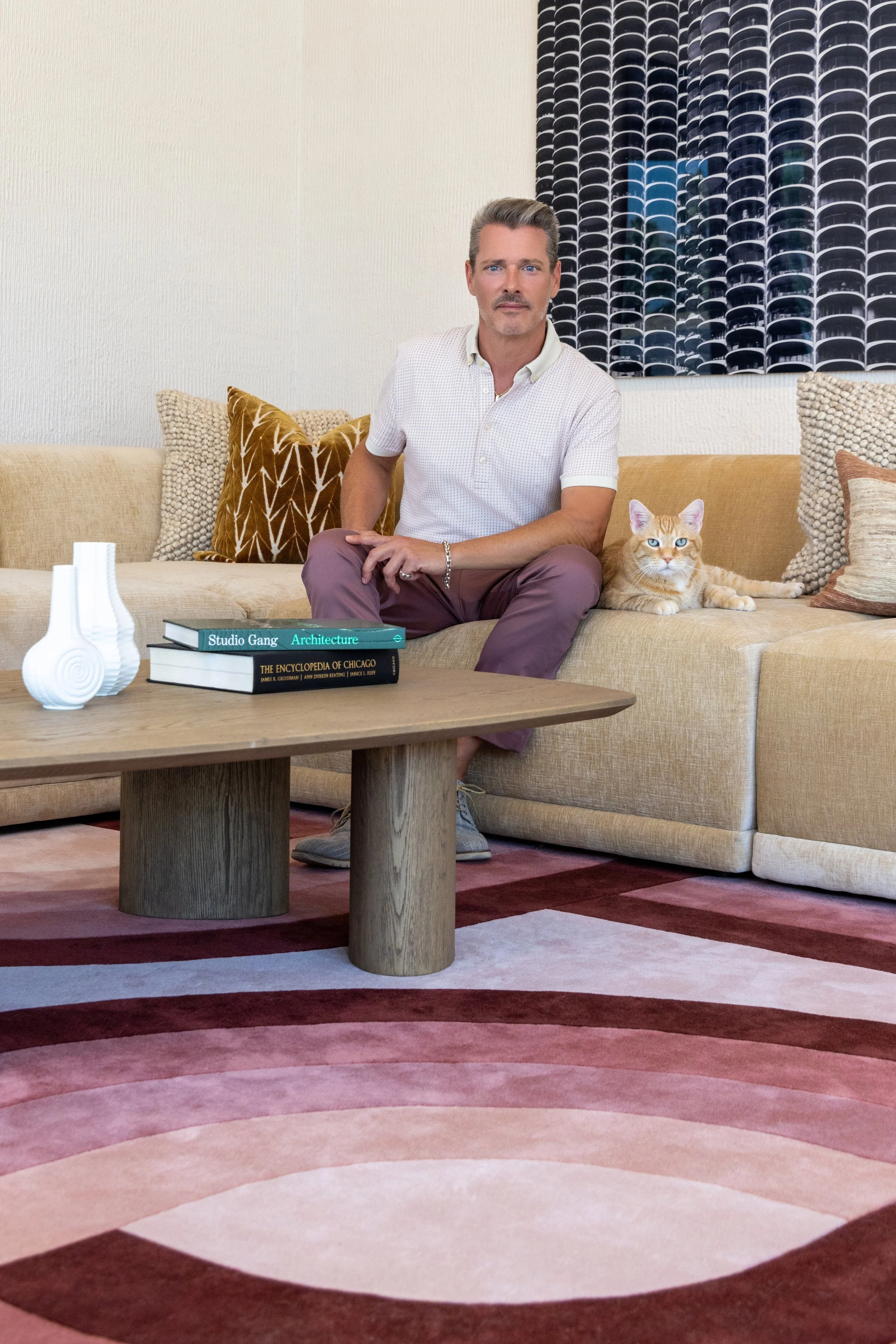 Man sitting on beige sofa with cat, decorative pillows, modern wooden coffee table, books, abstract rug, and artwork on wall.
