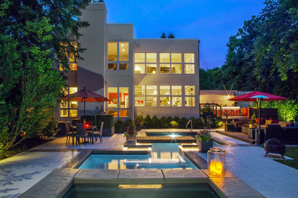 Modern house with large windows, a swimming pool, and patio area with furniture and umbrellas at night, illuminated by lights.