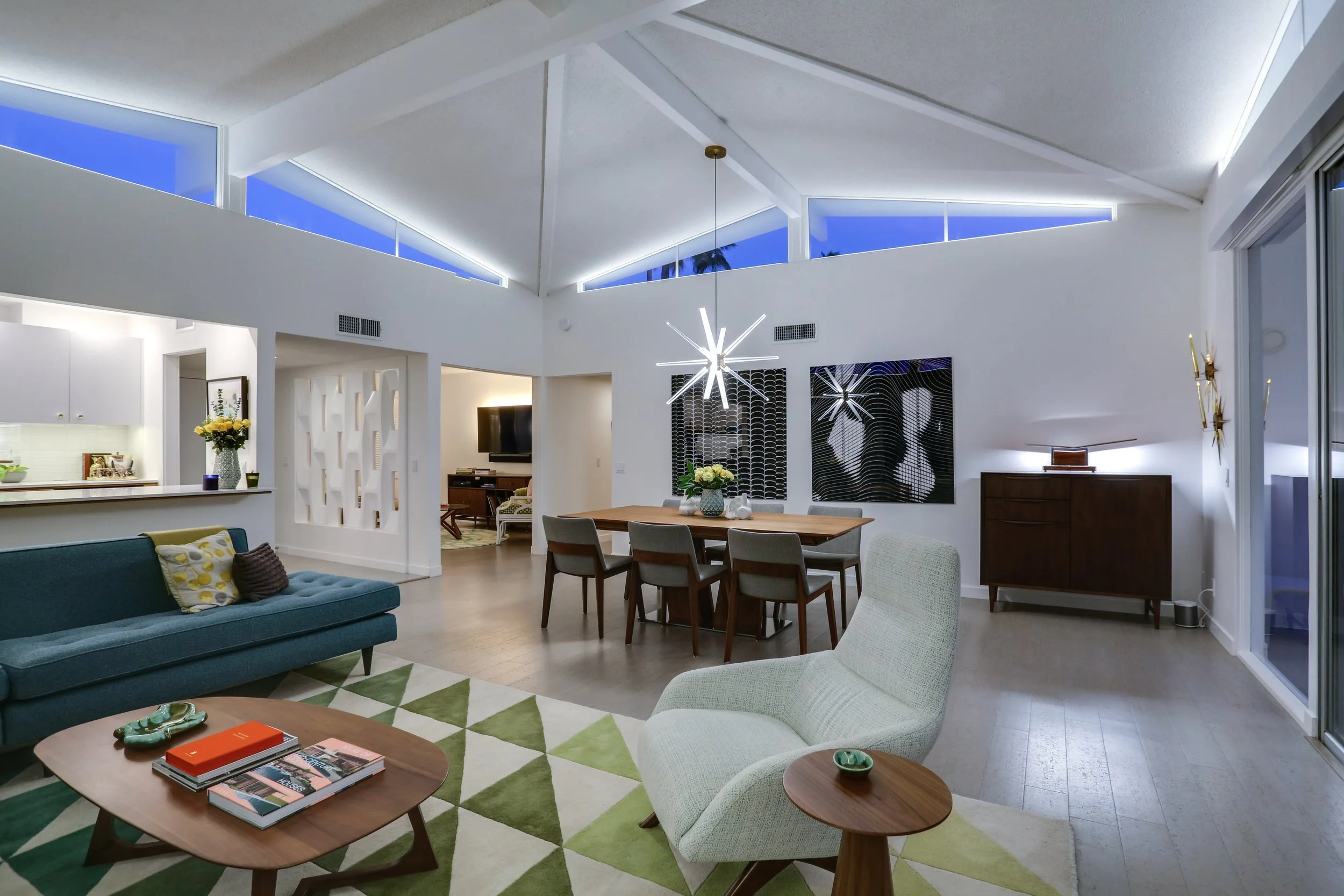 Modern living room with vaulted ceiling, featuring a blue sofa, geometric rug, and wooden coffee table. Contains a dining area with a large table, chairs, and abstract wall art. The room has large windows and contemporary lighting.