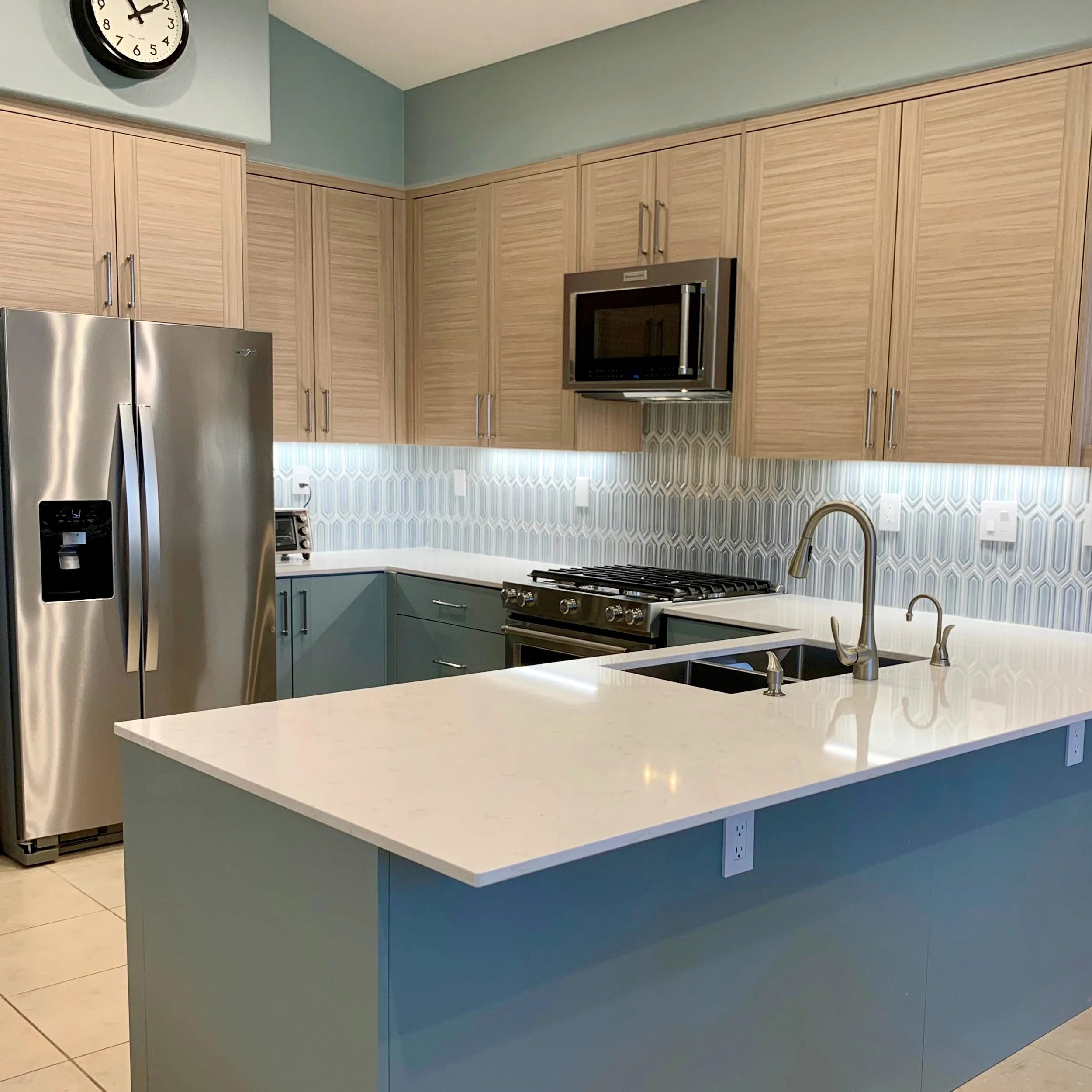 Modern kitchen with stainless steel appliances, light wood cabinets, white solid surface countertops, a geometric tile backsplash, and a wall clock.