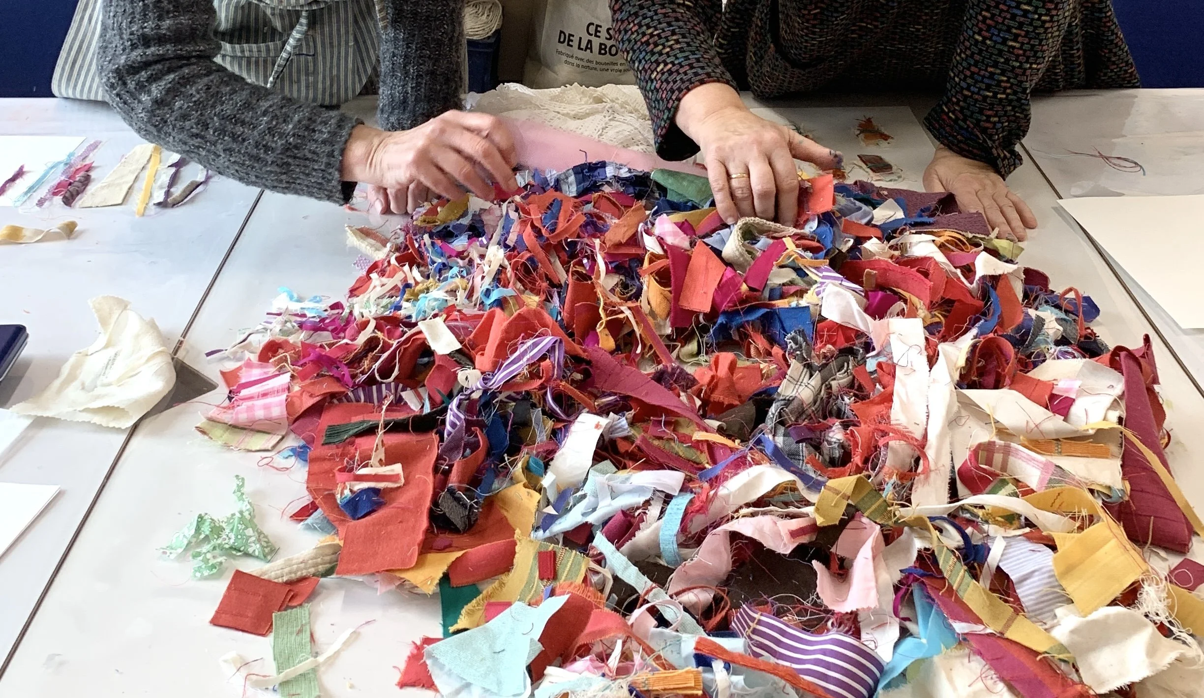 two people working with a large pile of colourful fabric scraps on a atble, hands sorting and selecting pieces in a workshop setting