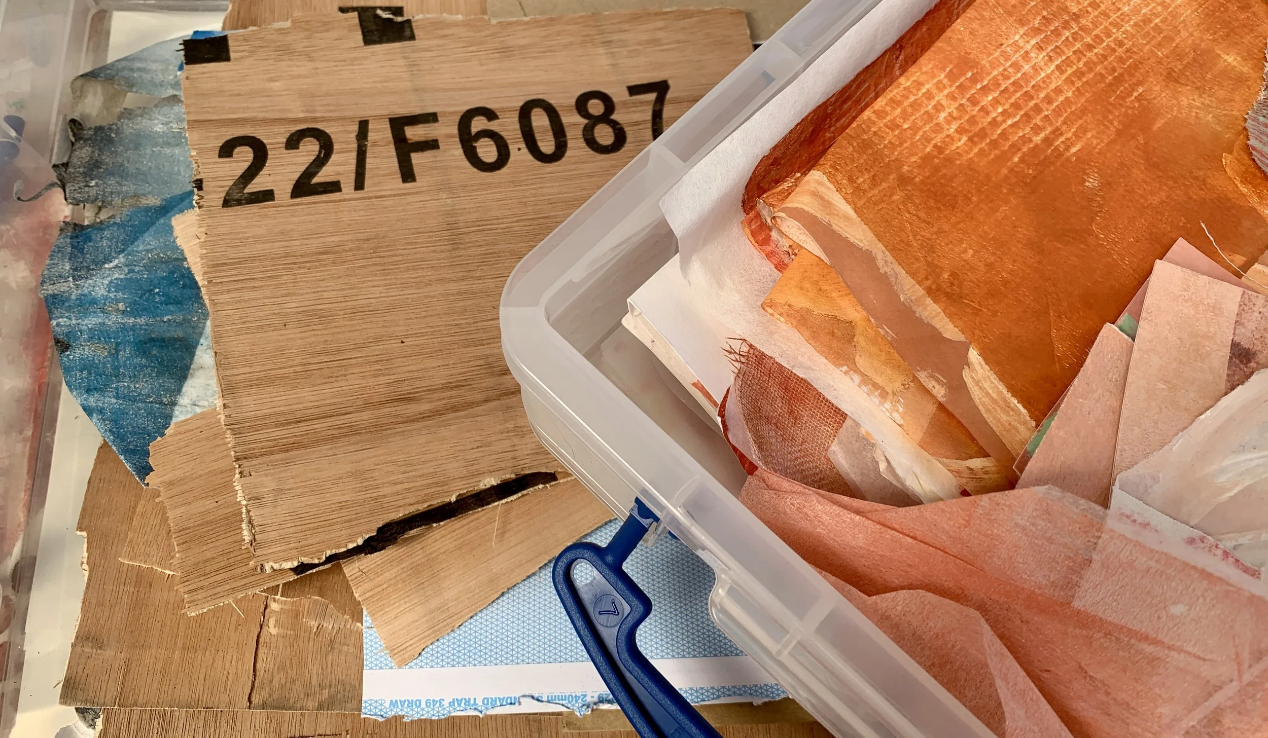Studio table with stacked wood offcuts, a plastic container filled with torn orange and peach painted papers, and layered collage materials in warm earthy tones.