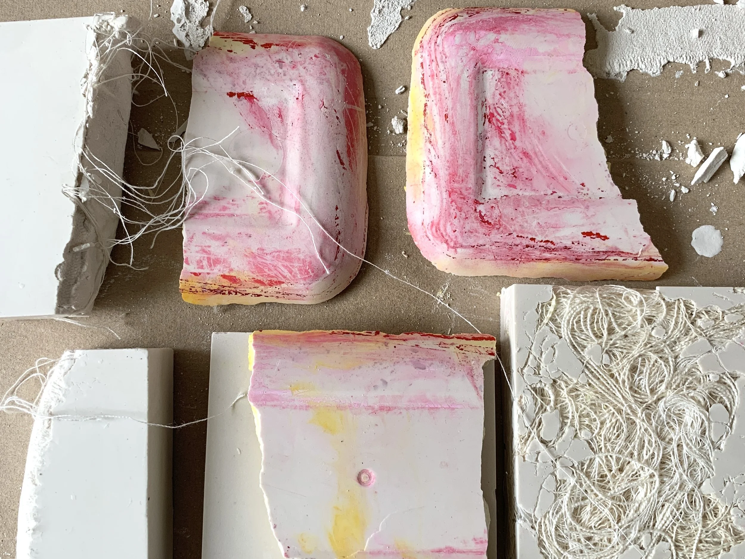 white plaster blocks arranged on a studio surface, some marked with soft pink and yellow pigment and embedded threads showing experimental surface texture