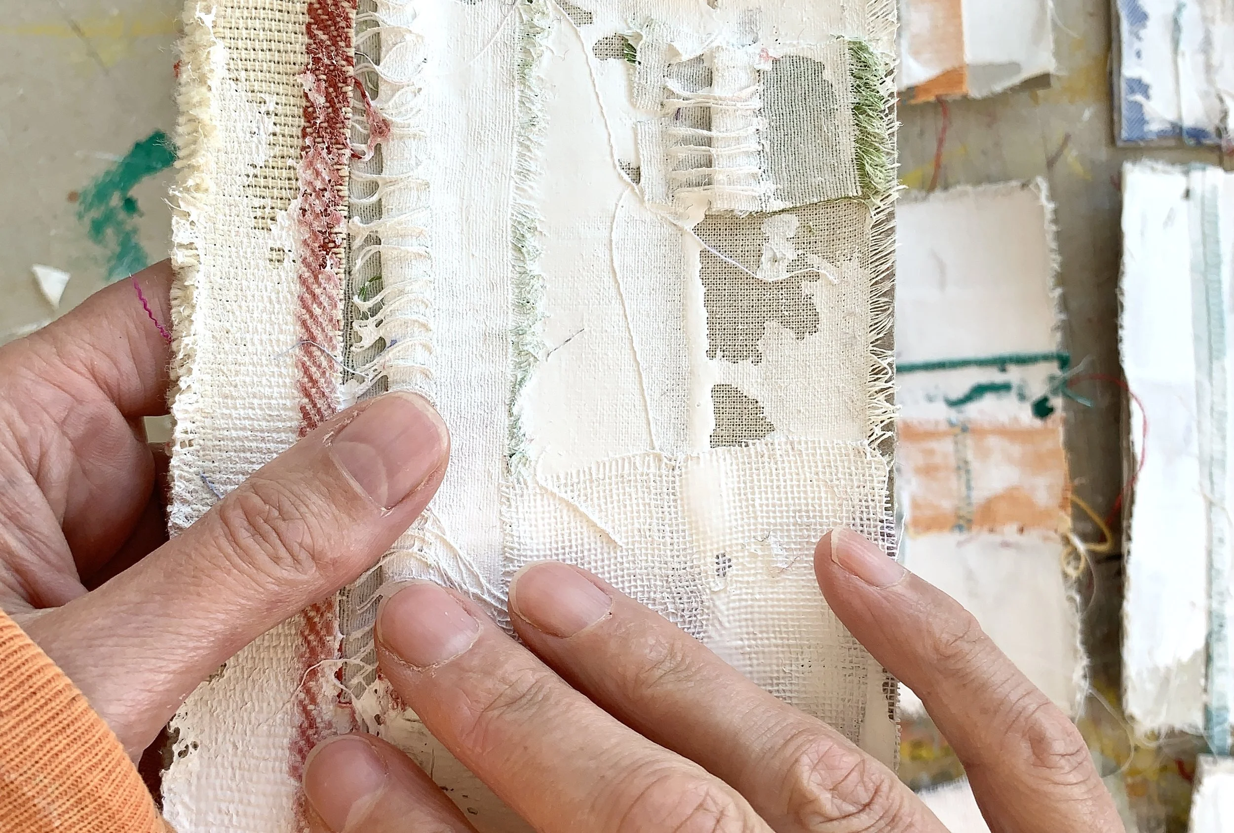 Hands holding a small textile collage made from layered fabric and paint, with frayed edges and stitched details. In the background other samll experimenatl pieces rest on a paint-marked studio surface, suggesting an ongoing process of making and exp