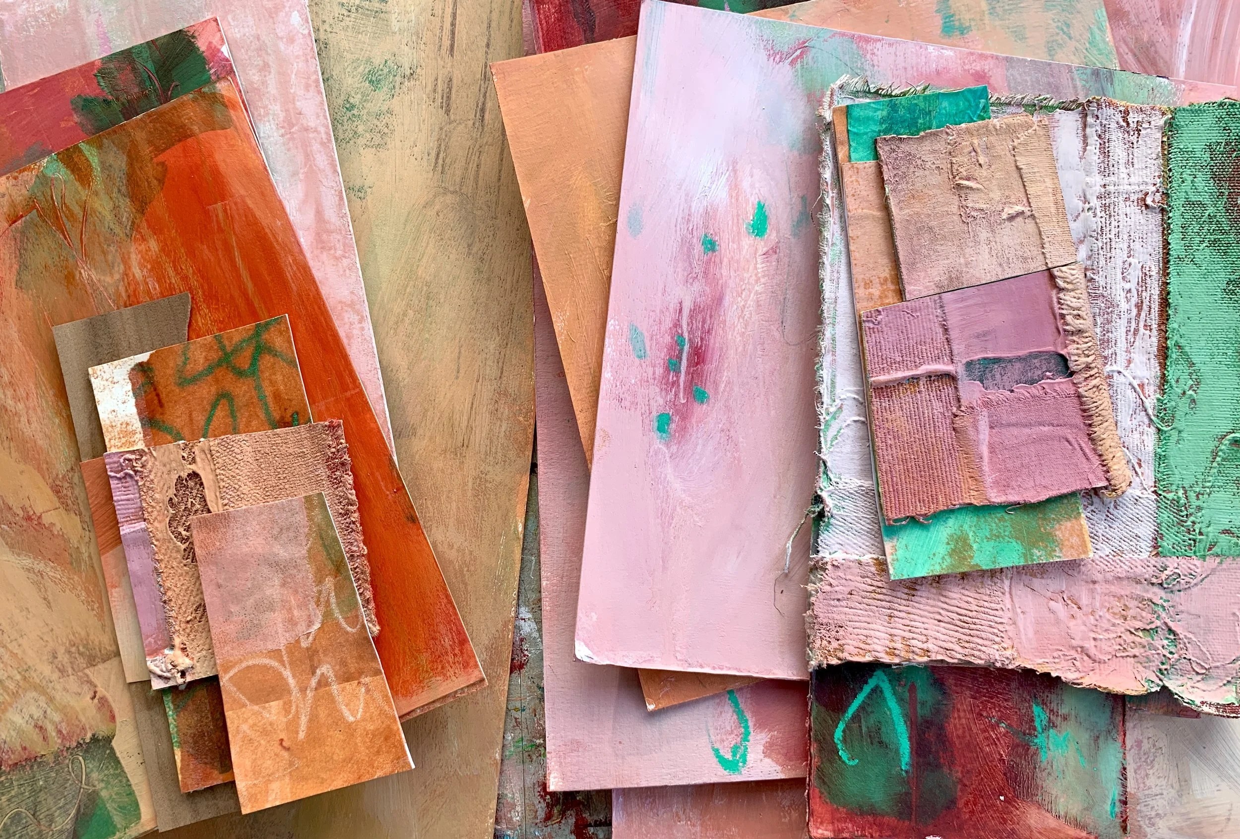 a collection of layered abstract collage pieces arranged on a studio table, combining painted card, fabric an textured surfaces in soft peach, pink and green tones. The works overlap and vary in size, showing visisble marks, edges and unrefined stage