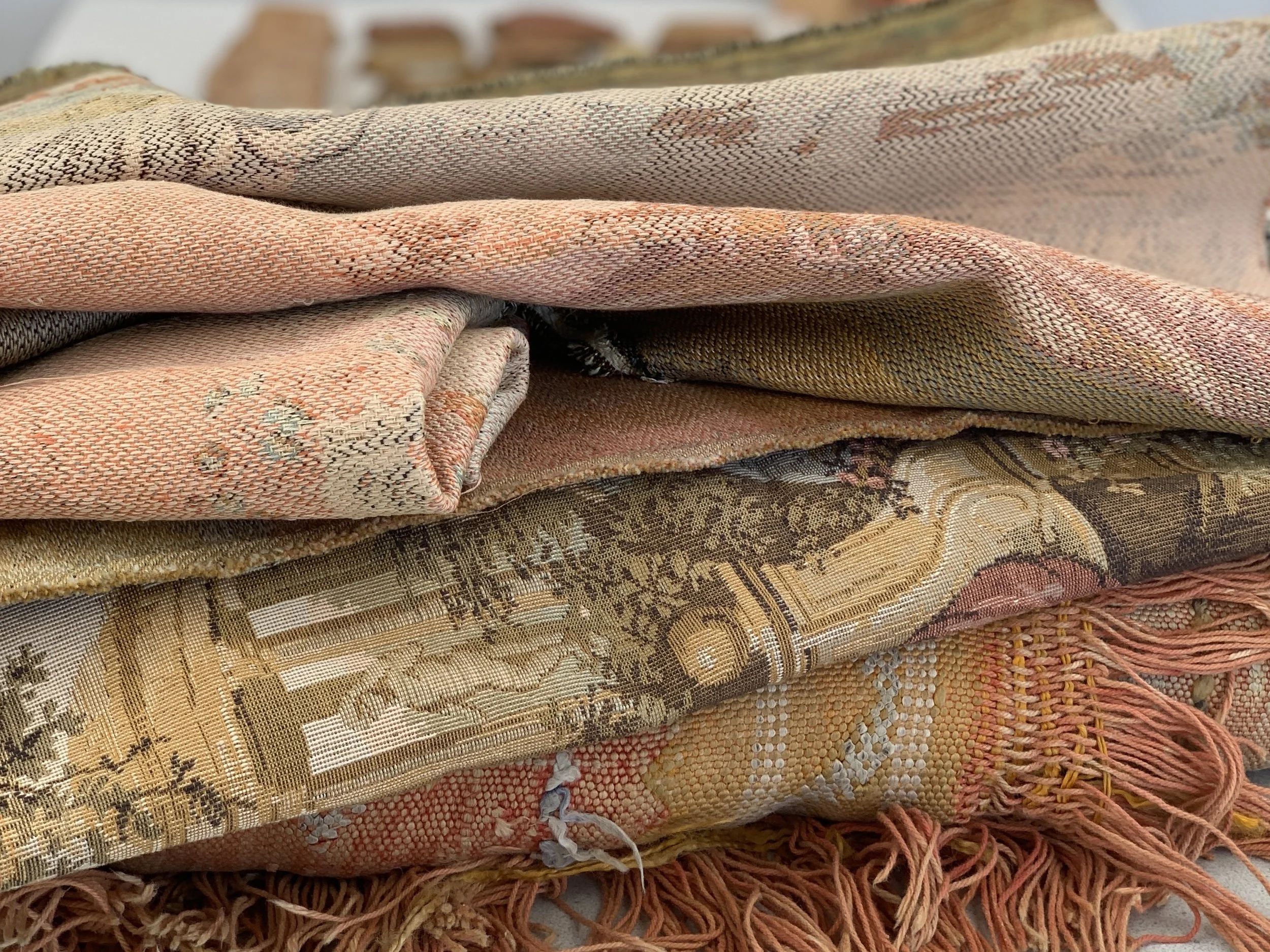 Folded woven fabric in soft ochre, terracotta adnd muted green tones with visible weave texture and frayed fringe.