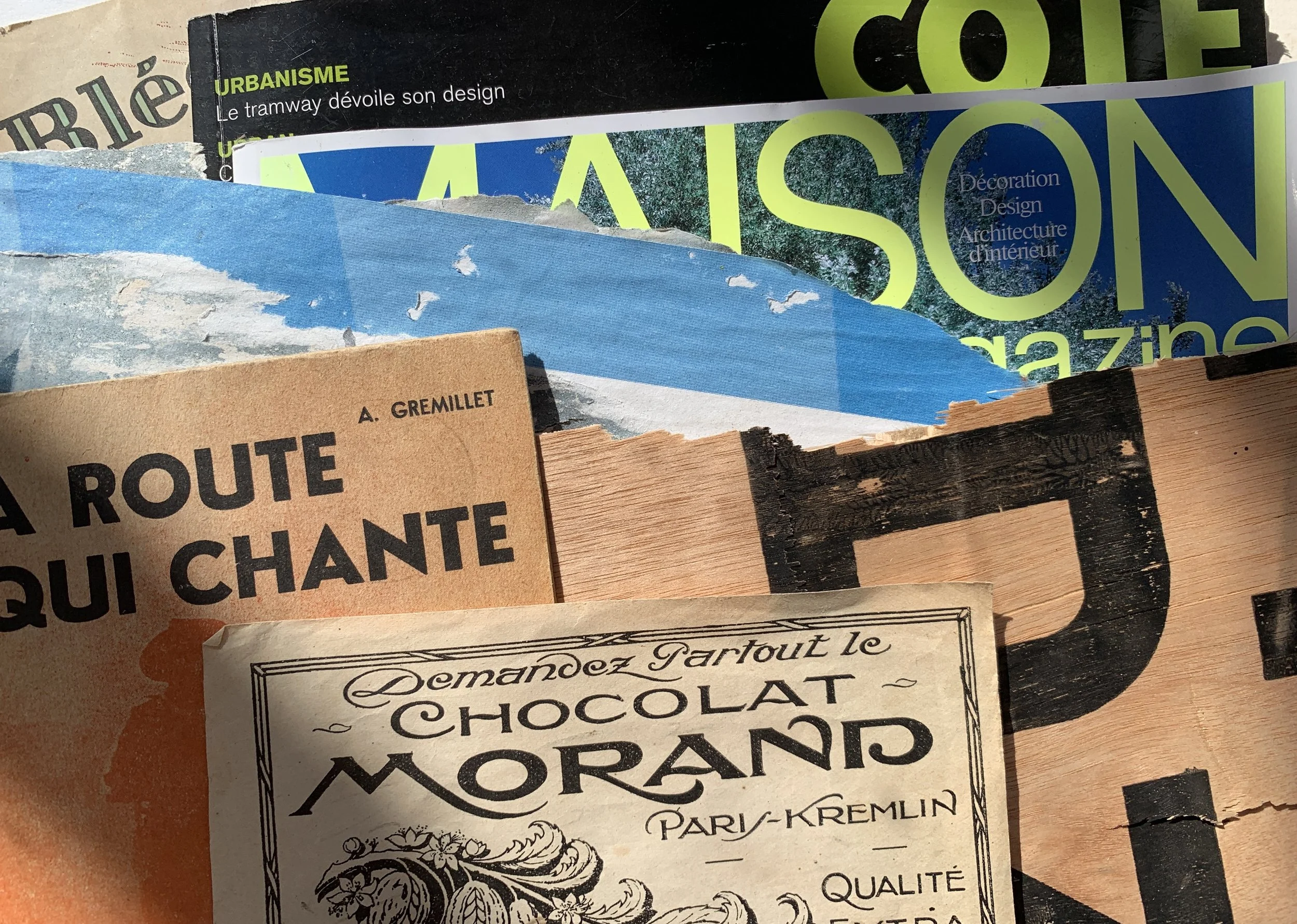 Layered collage materials including vintage French papers, a wood offcut with black lettering, torn magazine pages and a strip of blue painted paper arranged in overlapping textures.