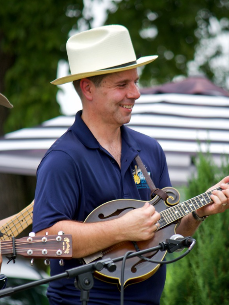 A man wearing a white hat and navy blue shirt playing a mandolin outdoors, with a striped umbrella and green trees in the background.