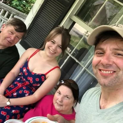 Four people smiling outdoors near a house with large windows, including a young woman in a red and blue dress, a man in a light shirt and cap, an older woman in a pink shirt, and an older man in a black shirt.