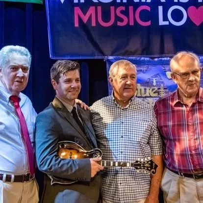 Four men standing together on a stage, with a large screen behind them displaying the words 'MUSIC' and 'LOVE' in colorful text.