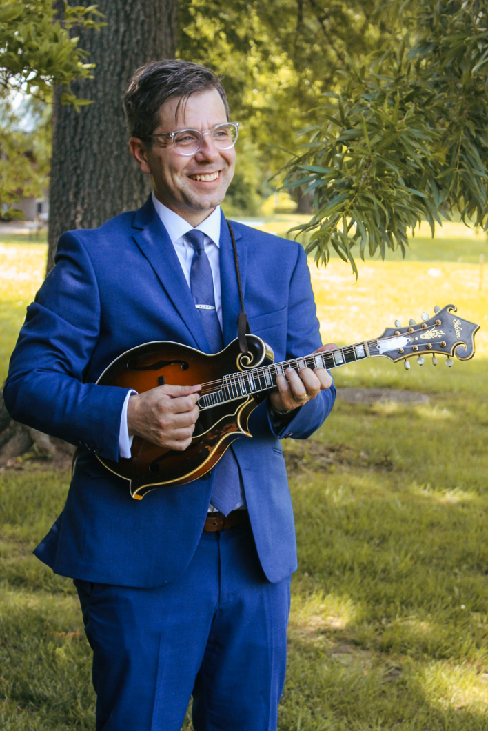 Man in a blue suit playing a mandolin outdoors in a park with green trees and grass.