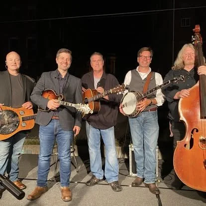 Five men standing on a stage holding musical instruments, including guitars, a banjo, and a double bass, with a dark background and stage equipment.