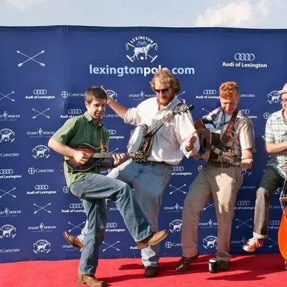 Four men playing musical instruments on a red carpet in front of a blue backdrop with the Lexington Police logo, Audi logos, and event sponsors.