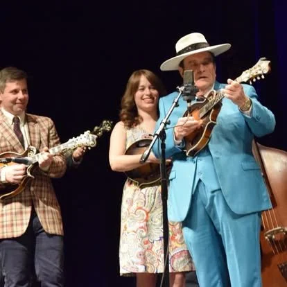 Three musicians performing on stage with guitars; a man in a blue suit and large hat, a woman in a colorful dress, and a man in a checkered jacket.