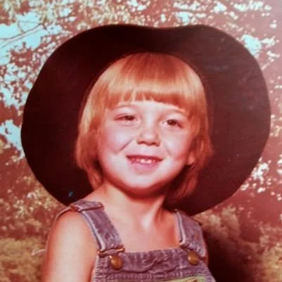 Young girl with blonde hair wearing a large black hat and denim overalls, smiling outdoors with trees in the background.