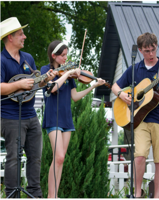 Three people performing music outdoors, playing a banjo, a violin, and a guitar, with a blue house and green trees in the background.