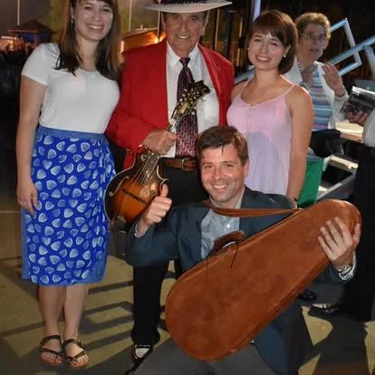 Group of five people at an outdoor event, with two men holding stringed instruments, and three women standing behind them, smiling.