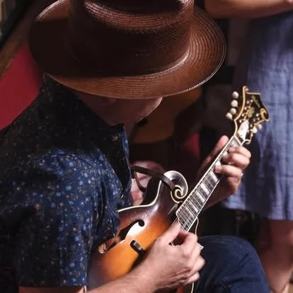 Person wearing a large straw hat, playing a mandolin in a dimly lit setting.