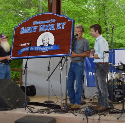 A man on stage at a Sandy Hook, Kentucky event, speaking into a microphone with two other men nearby. A large sign reads "Welcome to Sandy Hook KY" with a picture of a dog's face and mentions the home of the "Whizzy." Musical instruments and equipment are also on stage.