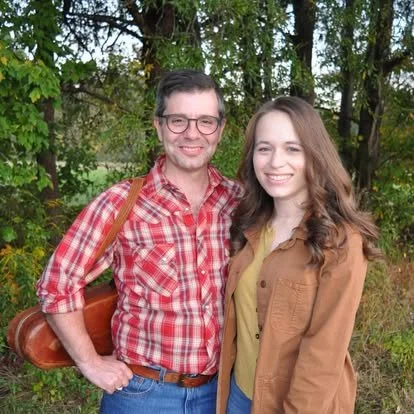 A man and a woman standing outdoors among trees, smiling at the camera. The man is wearing glasses, a red plaid shirt, and carrying a leather bag. The woman has long curly hair and is wearing a yellow top with a tan jacket.