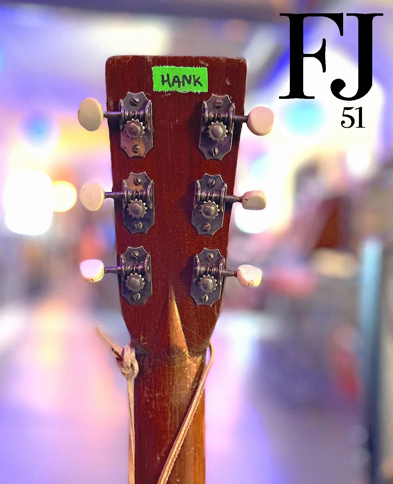 Close-up of the back of a guitar headstock with tuning pegs, with a green sticker labeled 'HANK' and blurred colorful background.