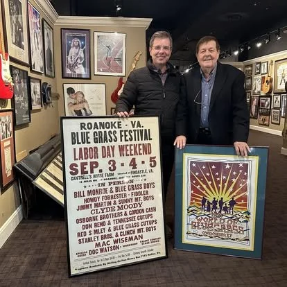 Two men standing indoors at an art gallery or museum, surrounded by framed artwork, holding a large poster advertising the Roanoke Virginia Blue Grass Festival for Labor Day Weekend, September 3-5, with details of performers and times.