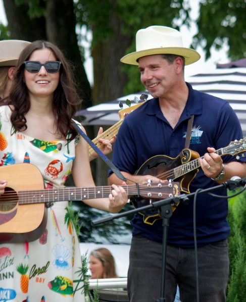 Two people playing acoustic guitars outdoors, smiling, with trees and tents in the background.