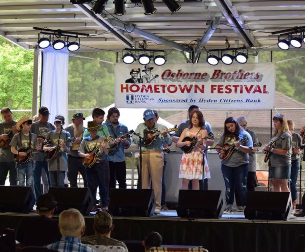 A group of people playing mandolins on a stage during the Oskorne Brothers Hometown Festival, sponsored by Hyden Citizens Bank, with an audience watching, outdoors with green trees in the background.