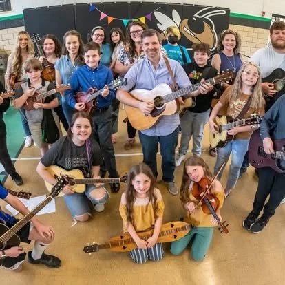 A group of children and a man holding guitars in a school gym, posing for a photo with musical instruments.