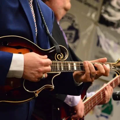 Person playing a stringed instrument, possibly a mandolin, wearing a blue jacket.