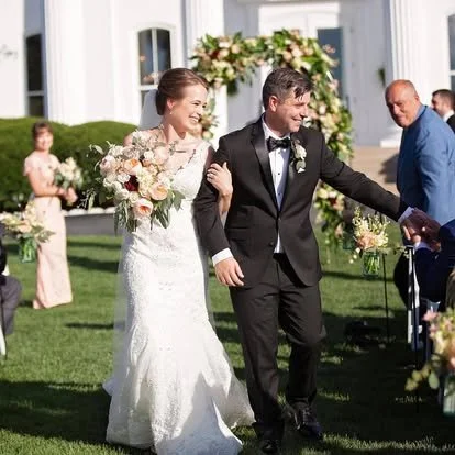 Bride and groom walking outdoors after wedding ceremony, smiling and holding hands, with wedding guests and floral arch in the background