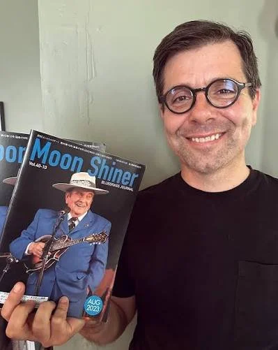 A man with glasses and a black shirt smiling while holding a magazine titled 'Moon Shiner' featuring an older man in a blue suit and hat playing a guitar on the cover.