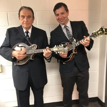 Two men in formal suits playing mandolins and smiling, standing against a plain white wall.
