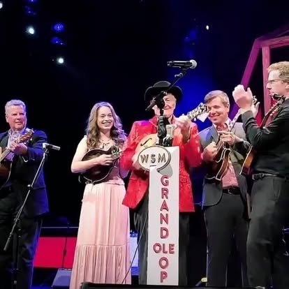 Musicians performing on stage at a grand ole opry event, with a woman in a pink dress, a man in a red jacket, and others playing instruments.