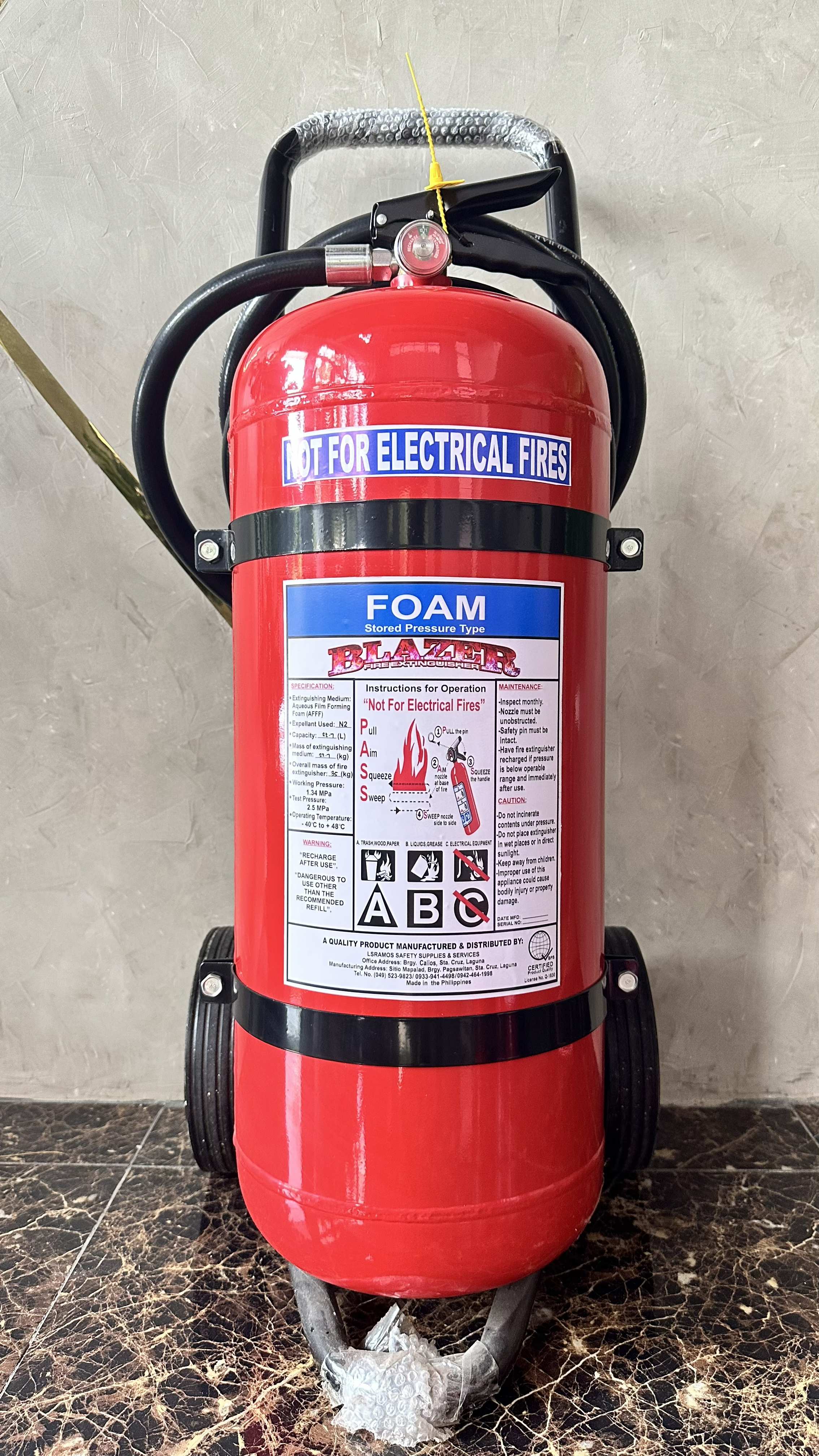 A red fire extinguisher with a label indicating it is for foam fires. The extinguisher has a yellow safety pin and yellow tube on top. It is placed on a black stand on a marble floor with a textured beige wall in the background.