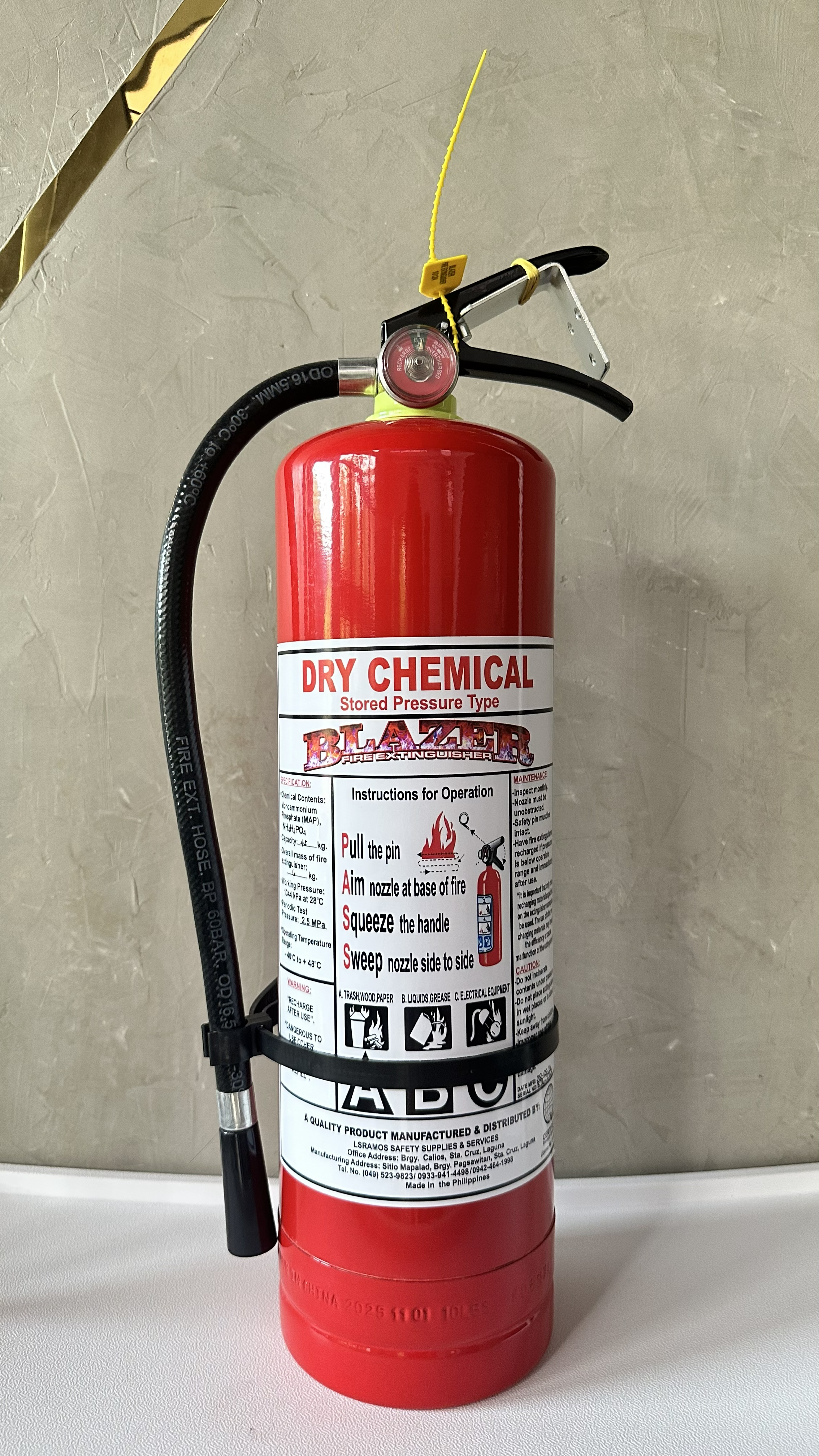 Red dry chemical fire extinguisher with instructions on the label and a black hose attached, placed on a white surface against a beige wall.