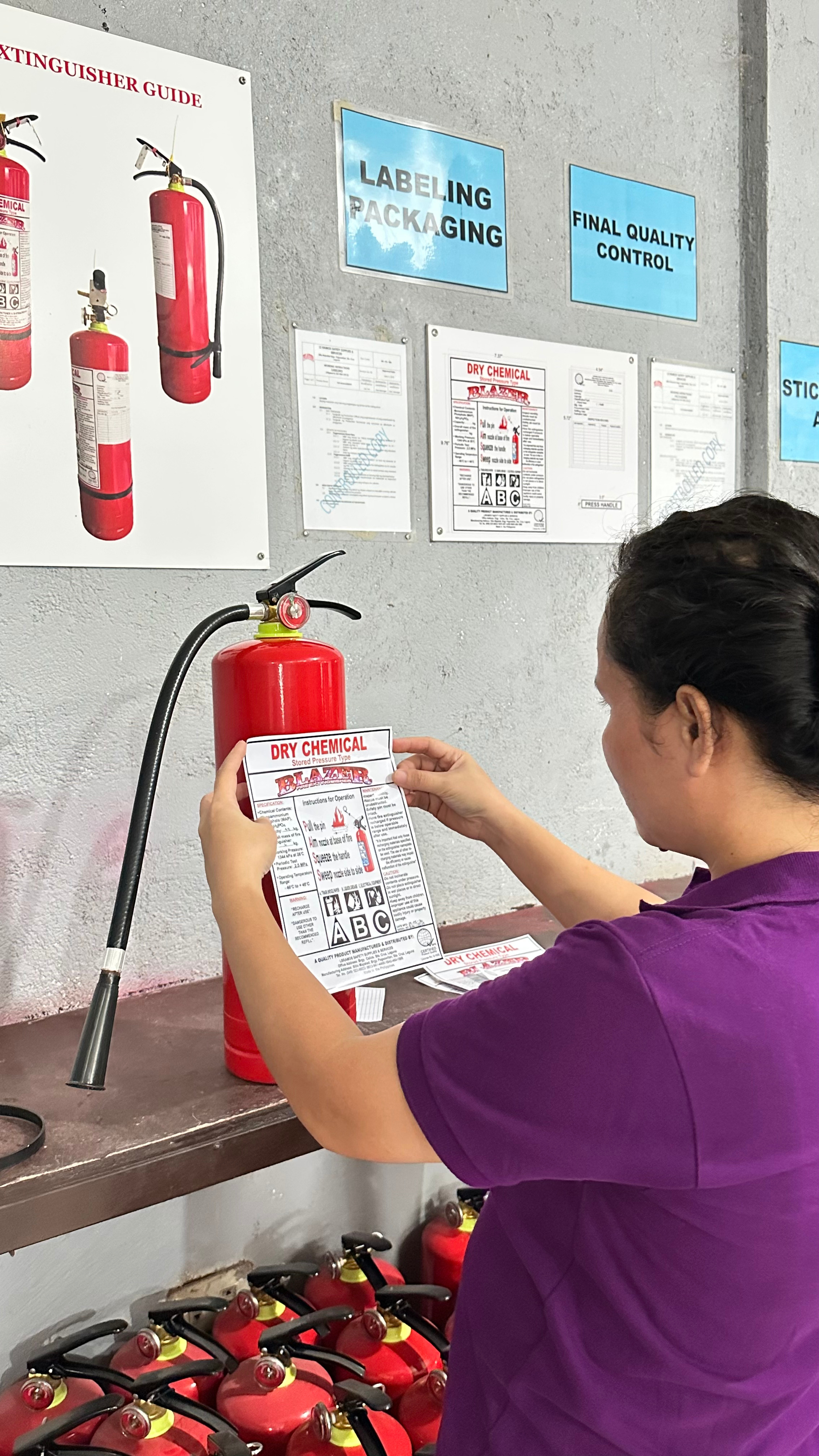 A person wearing a purple shirt is holding a fire extinguisher with a label that says 'Dry Chemical.' They are in a room with multiple fire extinguishers on a table and on the floor, with safety and labeling signs on the wall behind them. The signs include 'Labeling Packaging' and 'Final Quality Control.'