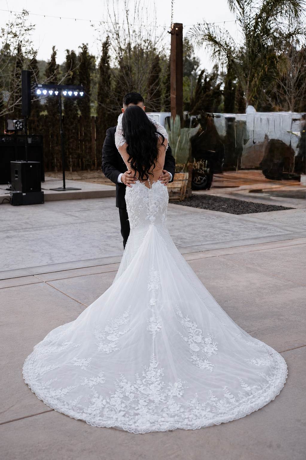 A bride and groom are dancing outdoors at sunset. The bride is in a white wedding gown with a long train, and the groom is in a black tuxedo.