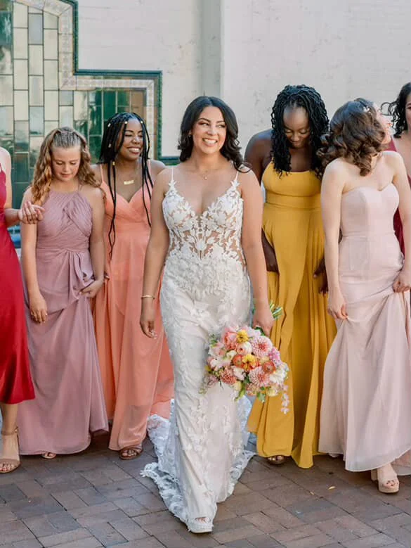 A bride in a white lace wedding gown holding a bouquet, surrounded by bridesmaids in pastel-colored dresses, smiling outdoors.