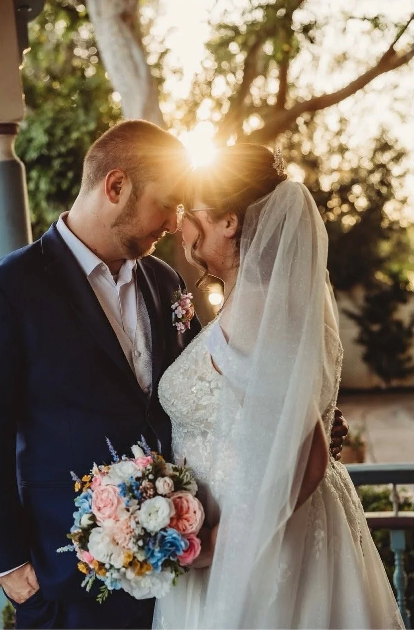A bride and groom share a tender moment at sunset outdoors, with the sun setting behind them. The groom holds a colorful bouquet of flowers, and the bride wears a lace wedding gown with a veil.