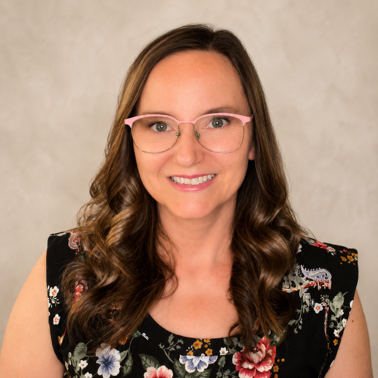 A woman with long wavy brown hair, wearing pink glasses and a black floral sleeveless top, smiling at the camera against a neutral background.