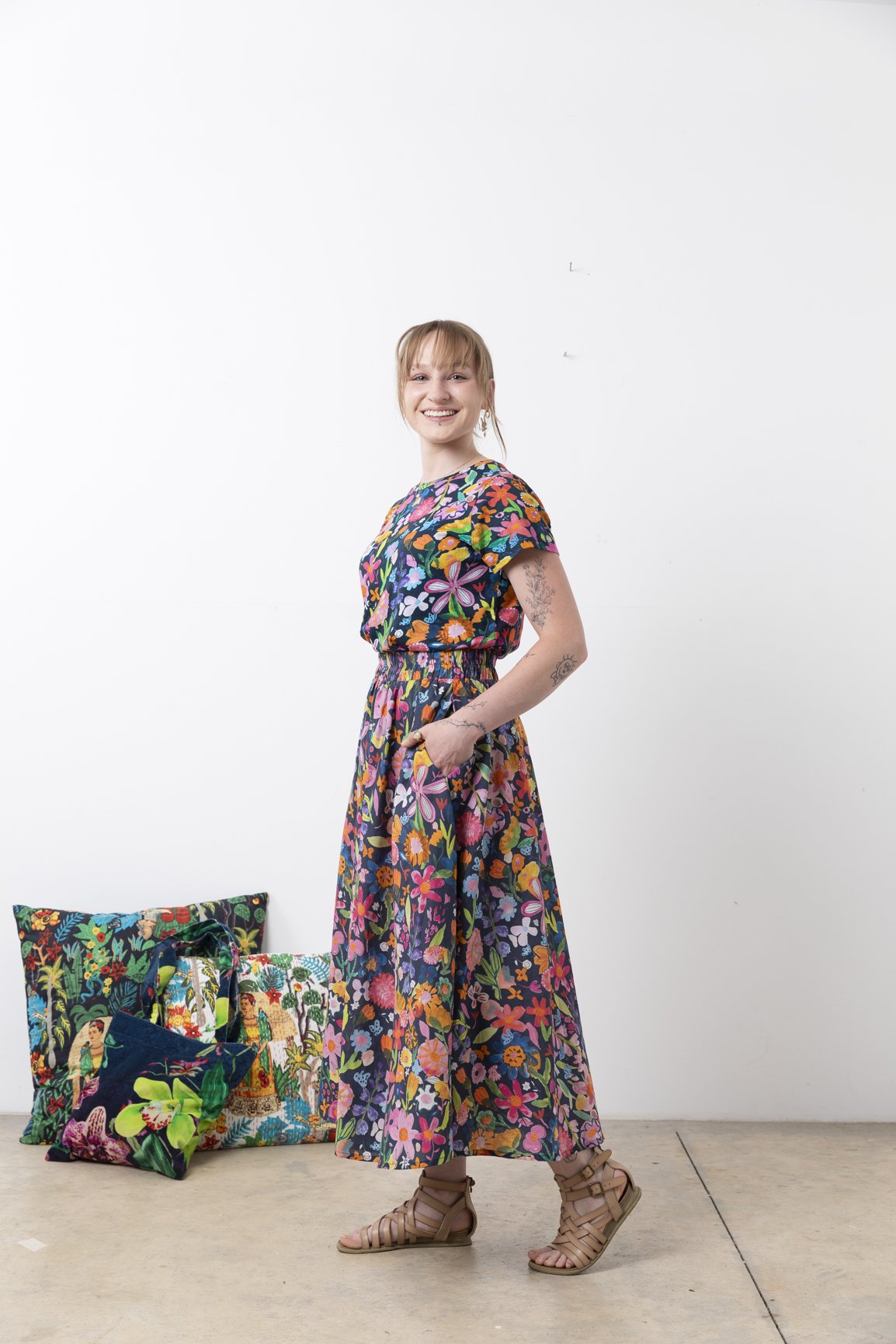 A young woman with blonde hair, wearing a colorful floral dress and sandals, stands against a white wall with her hands in her pockets, smiling. There are four cushions with vibrant, patterned designs on the floor beside her.