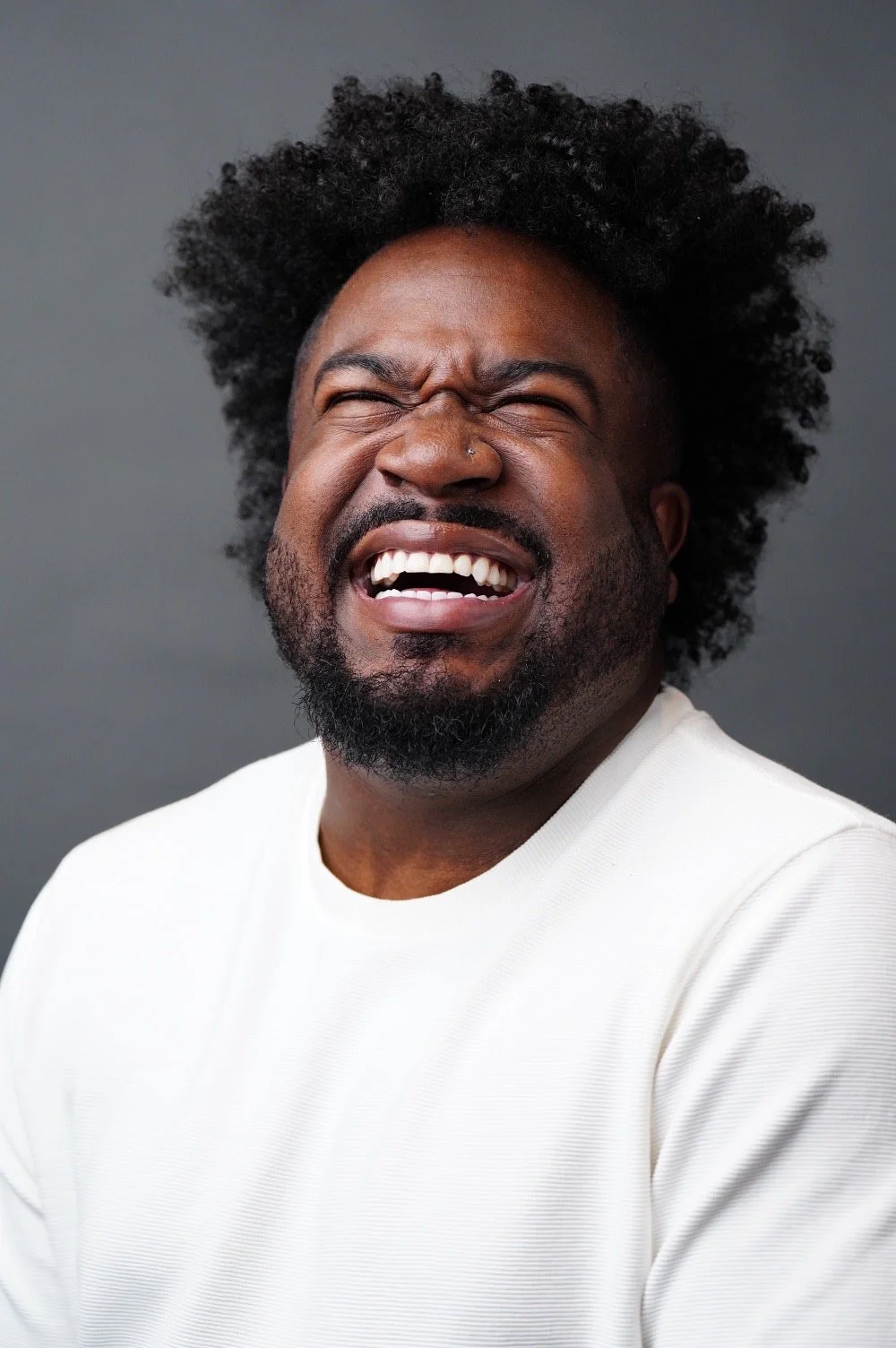 Man with curly black hair and beard laughing with eyes closed against a gray background.